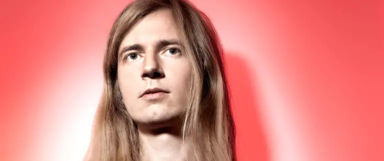 A young music producer with long hair in front of a red background.