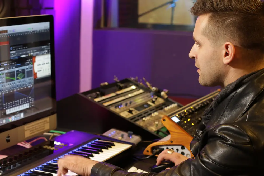 A man in a leather jacket is beatmaking on a keyboard in front of a monitor.