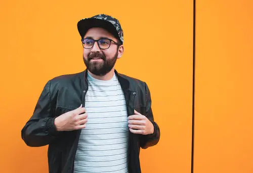 A bearded man in a black jacket and striped t-shirt standing in front of an orange wall, showcasing his music producer program.